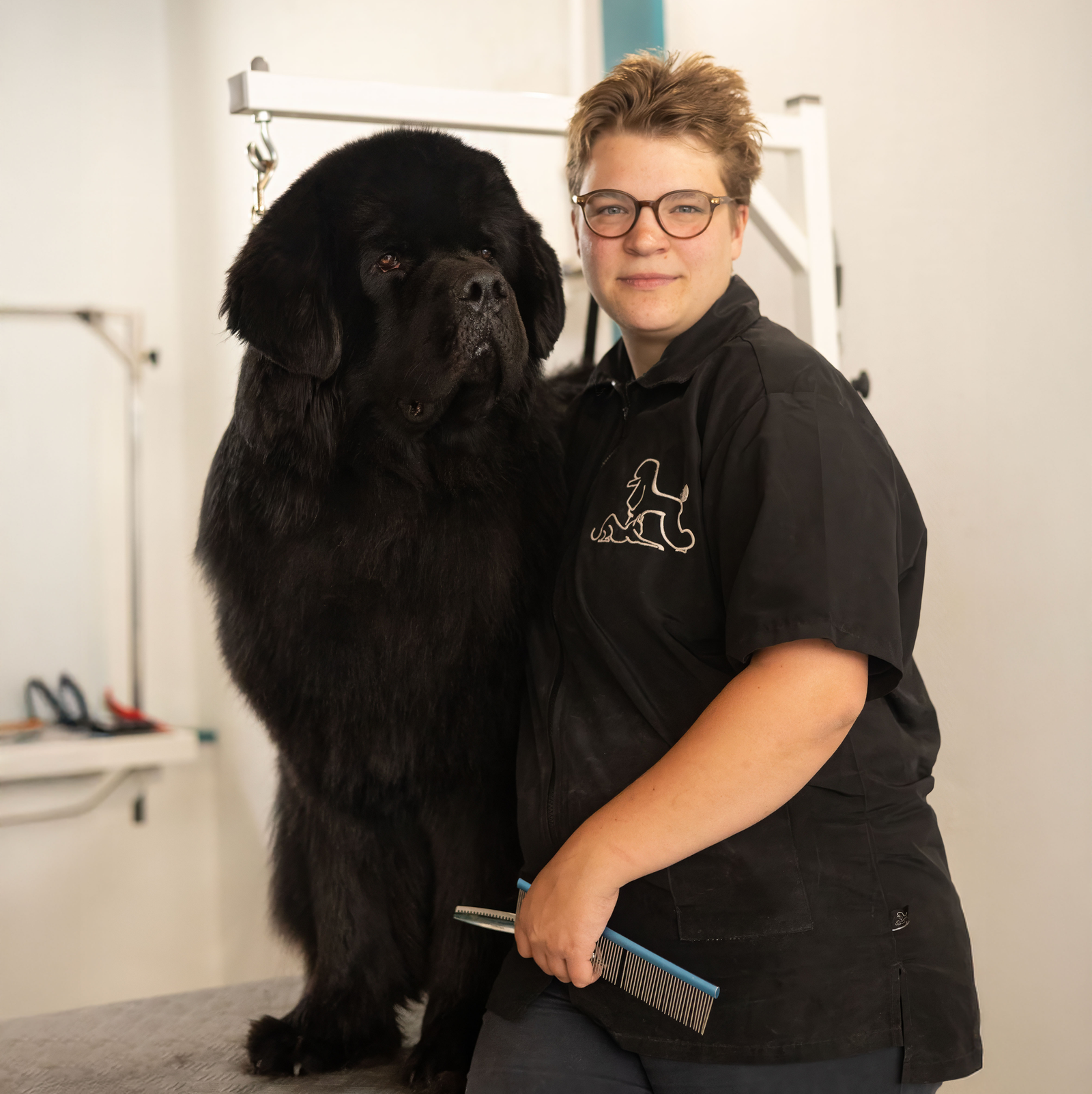 Toiletteuse souriante posant avec un grand chien de race Terre Neuve noir sur une table de toilettage.